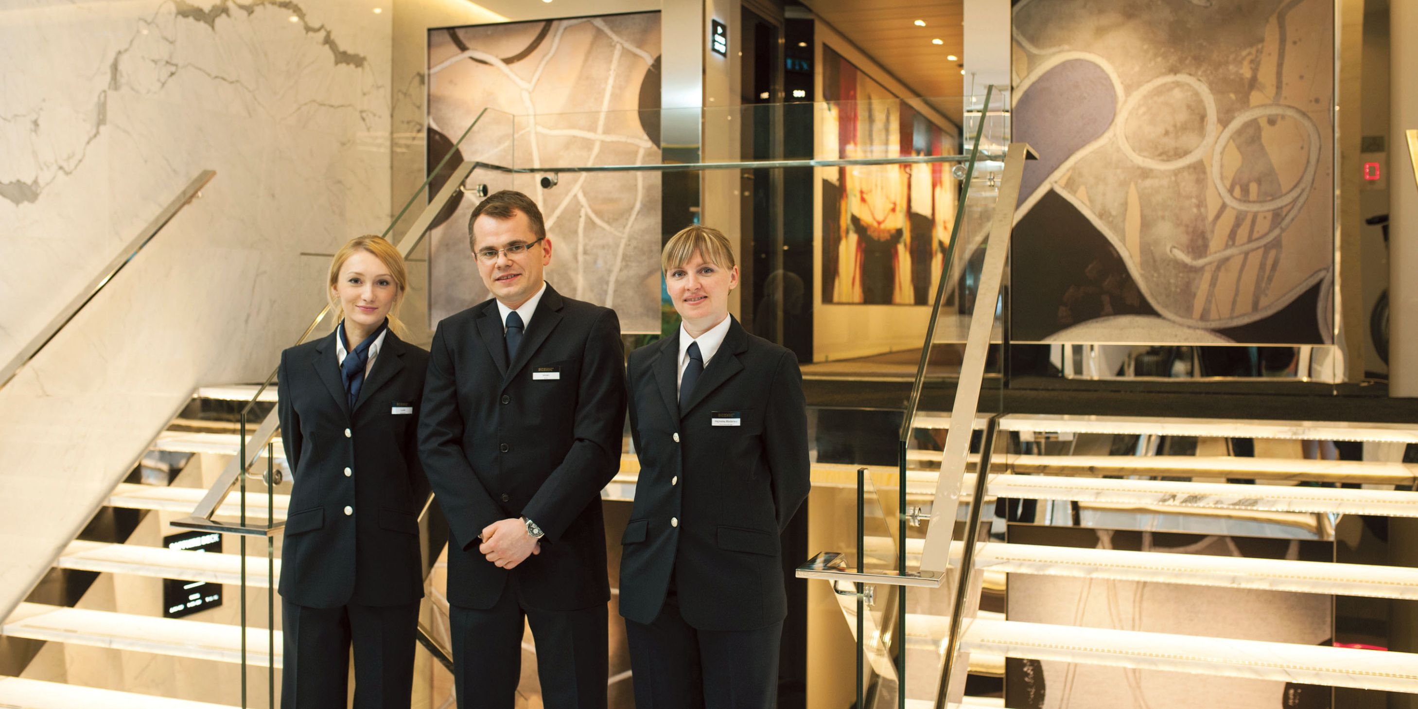 three crew members dressed in smart uniforms standing and smiling in front of a glass staircase