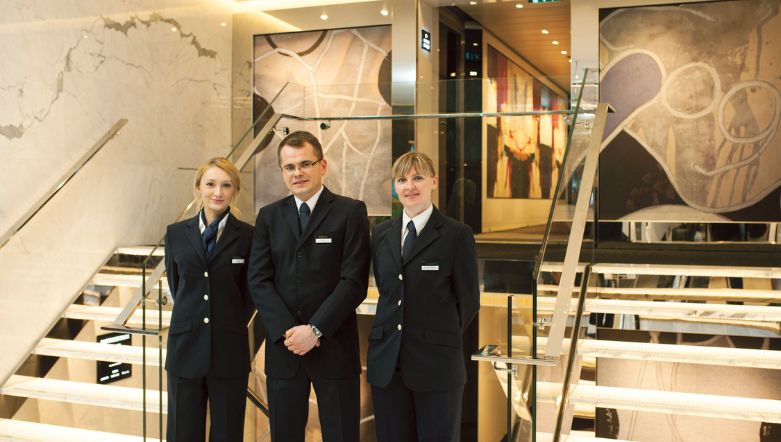 three crew members dressed in smart uniforms standing and smiling in front of a glass staircase